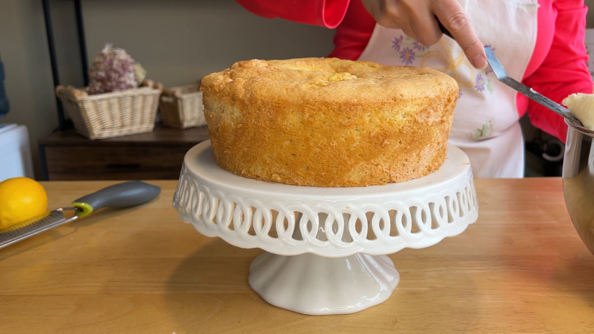 A person in a red sleeve prepares to frost a plain round cake on a white cake stand, with kitchen utensils and baskets in the background.
