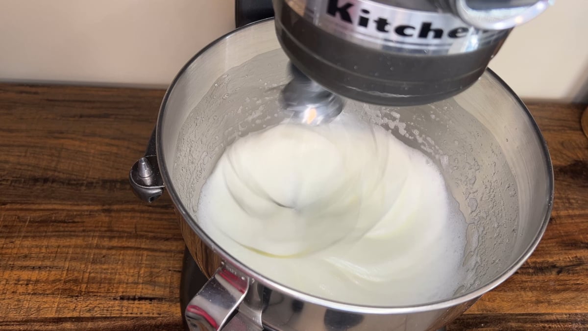 Electric stand mixer whipping egg whites in a stainless steel mixing bowl on a wooden countertop.