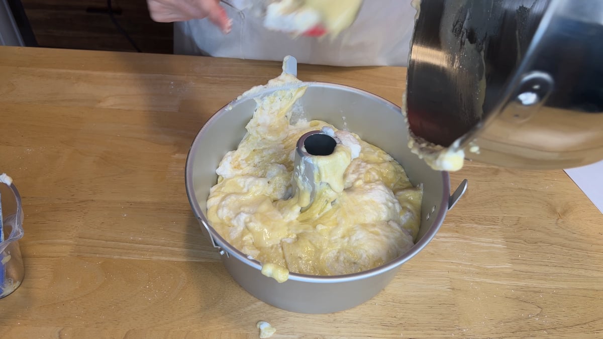 Cake batter being poured from a mixing bowl into a tube pan on a wooden surface, with some batter splattered on the pan and table.