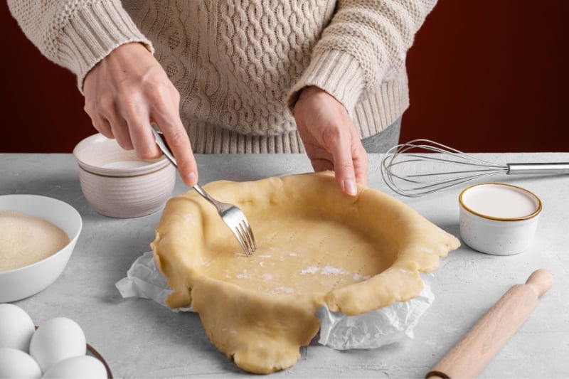 Person using a fork to prick holes in raw pie crust in a baking pan, with baking ingredients and utensils on the countertop.
