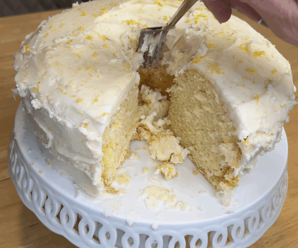 A partially sliced lemon cake with white frosting and lemon zest sits on a white cake stand; a hand holds a fork, taking a piece from the cake.