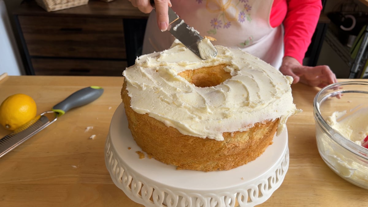 A person spreads white frosting on a round bundt cake placed on a white cake stand, with a bowl of frosting and a zester on the wooden table nearby.