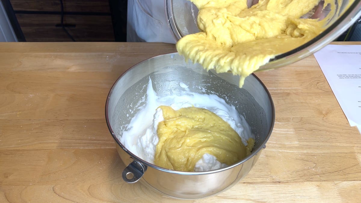 A bowl of yellow batter is being poured into a metal bowl containing whipped egg whites on a wooden surface.