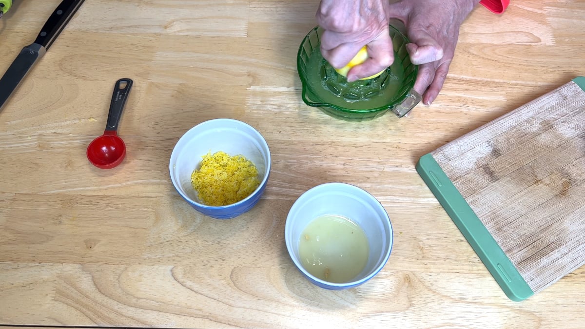 A person squeezes a lemon using a green glass juicer. Nearby are a bowl of lemon zest, a bowl of lemon juice, a red measuring spoon, a cutting board, and a knife on a wooden table.