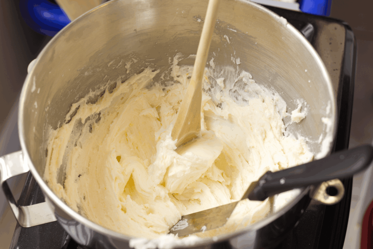 A metal mixing bowl containing creamy, whipped butter mixture with a wooden spoon and spatula resting inside.