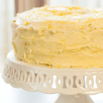 A frosted layer cake with a textured yellow and white icing sits on a white decorative cake stand.