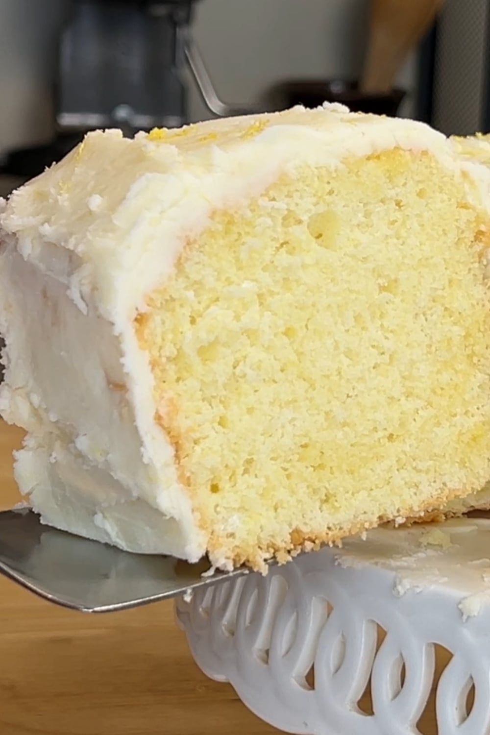 A close-up of a slice of lemon chiffon cake with white frosting being lifted from a decorative white cake stand.