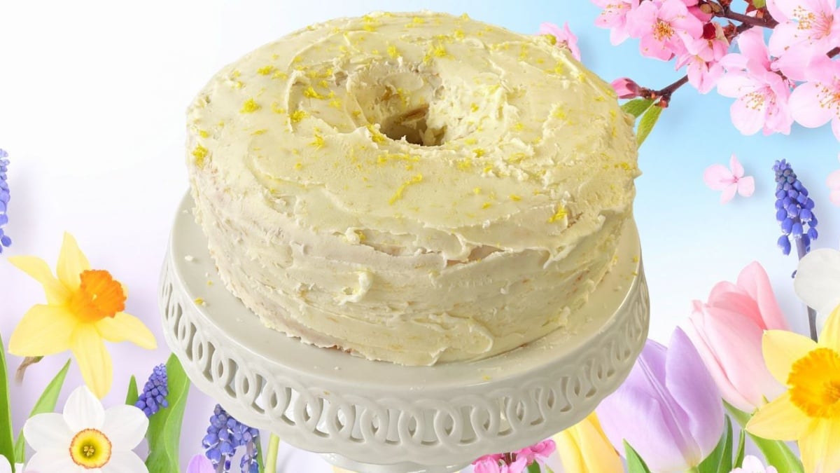 A lemon chiffon cake, frosted and topped with lemon zest, sits on a white cake stand surrounded by vibrant spring flowers like daffodils, tulips, and cherry blossoms.