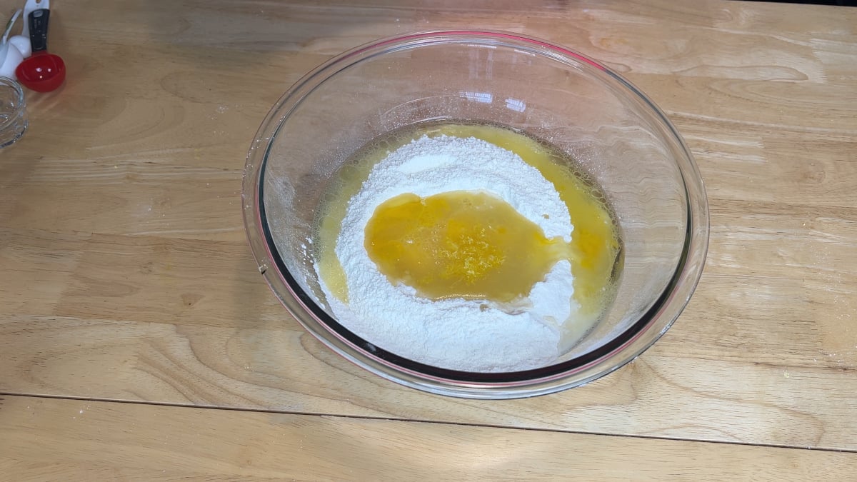 A glass bowl on a wooden table contains flour and a pool of beaten eggs, possibly for baking or making dough.