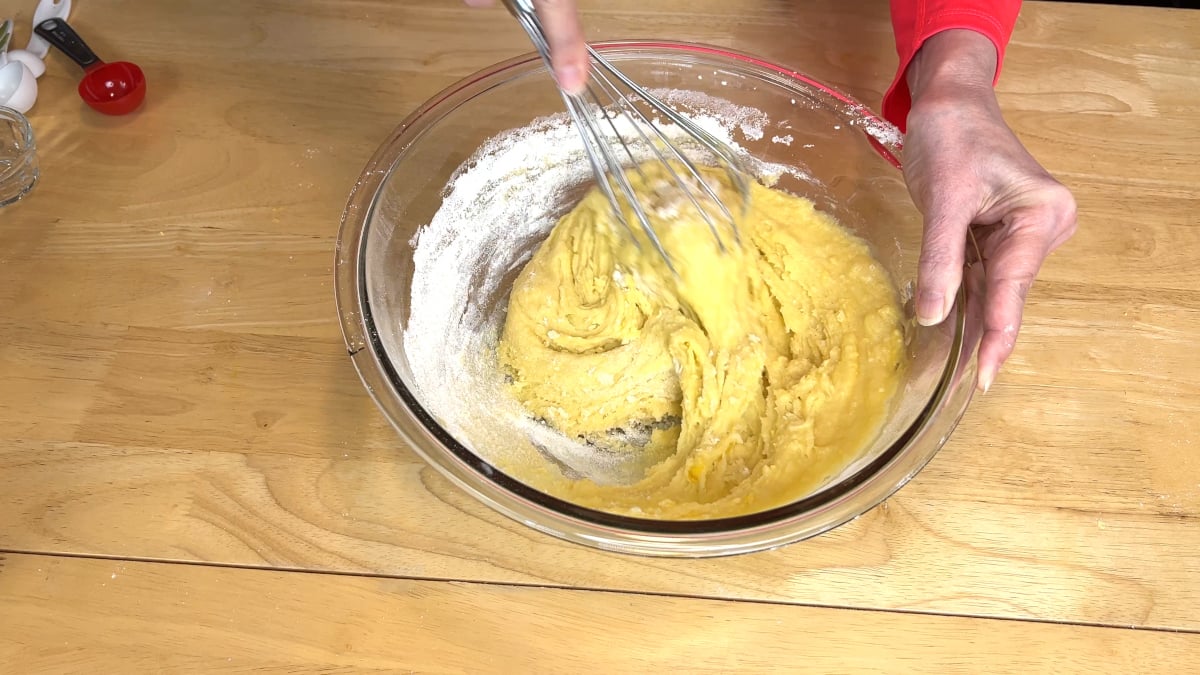A person whisking yellow batter in a glass bowl on a wooden table, with flour visible around the edges of the bowl.