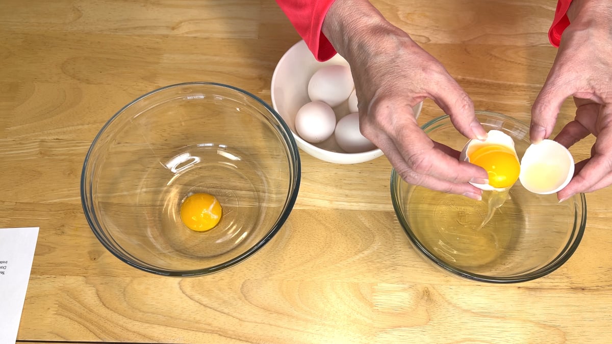 A person separates egg yolks from egg whites over two glass bowls, with a bowl of whole eggs nearby on a wooden table.