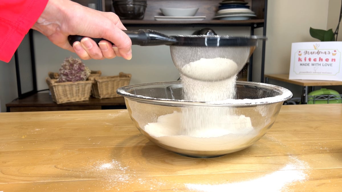 A hand sifting flour through a metal sieve into a glass mixing bowl on a wooden countertop.