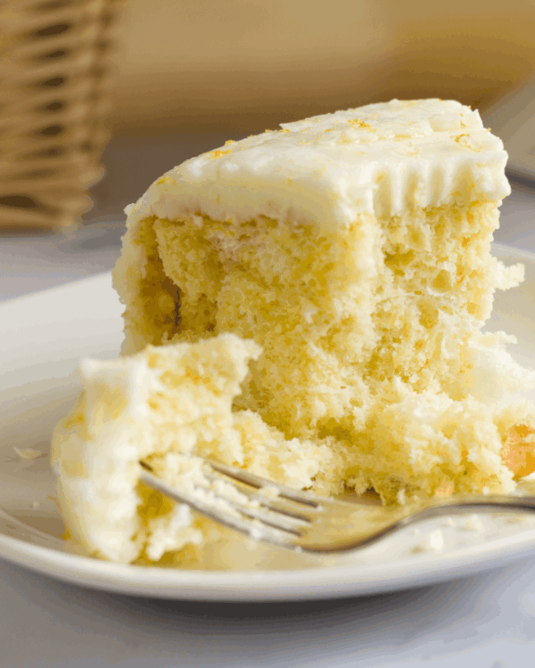 A close-up of a slice of yellow cake with white frosting on a plate, with a fork taking a bite from the piece.