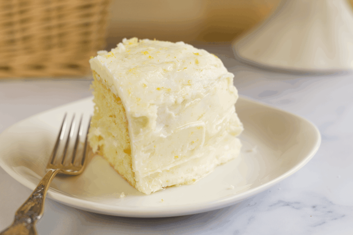 A slice of yellow cake with white frosting sits on a white plate next to a fork.