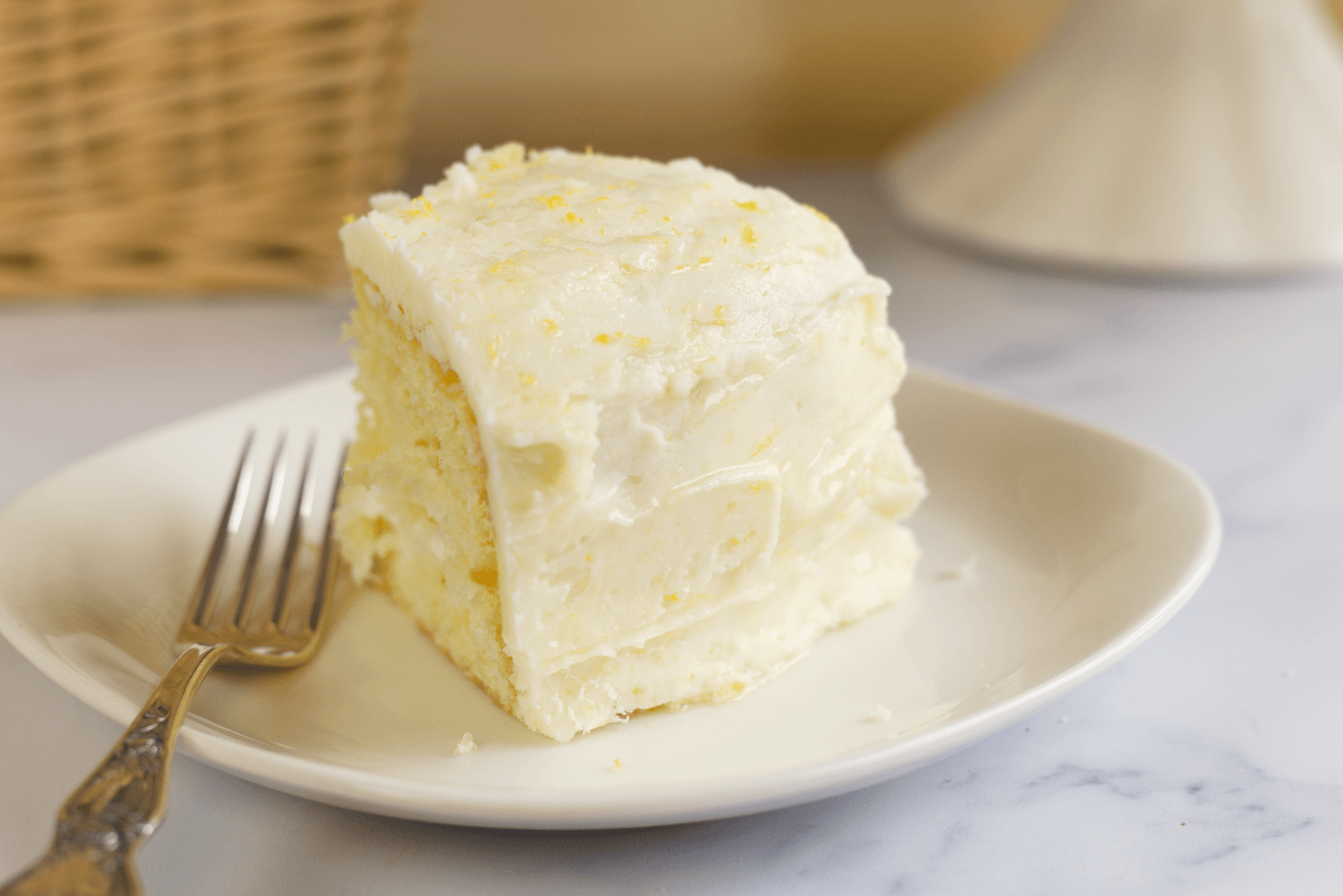 A slice of yellow cake with white frosting sits on a white plate next to a fork.