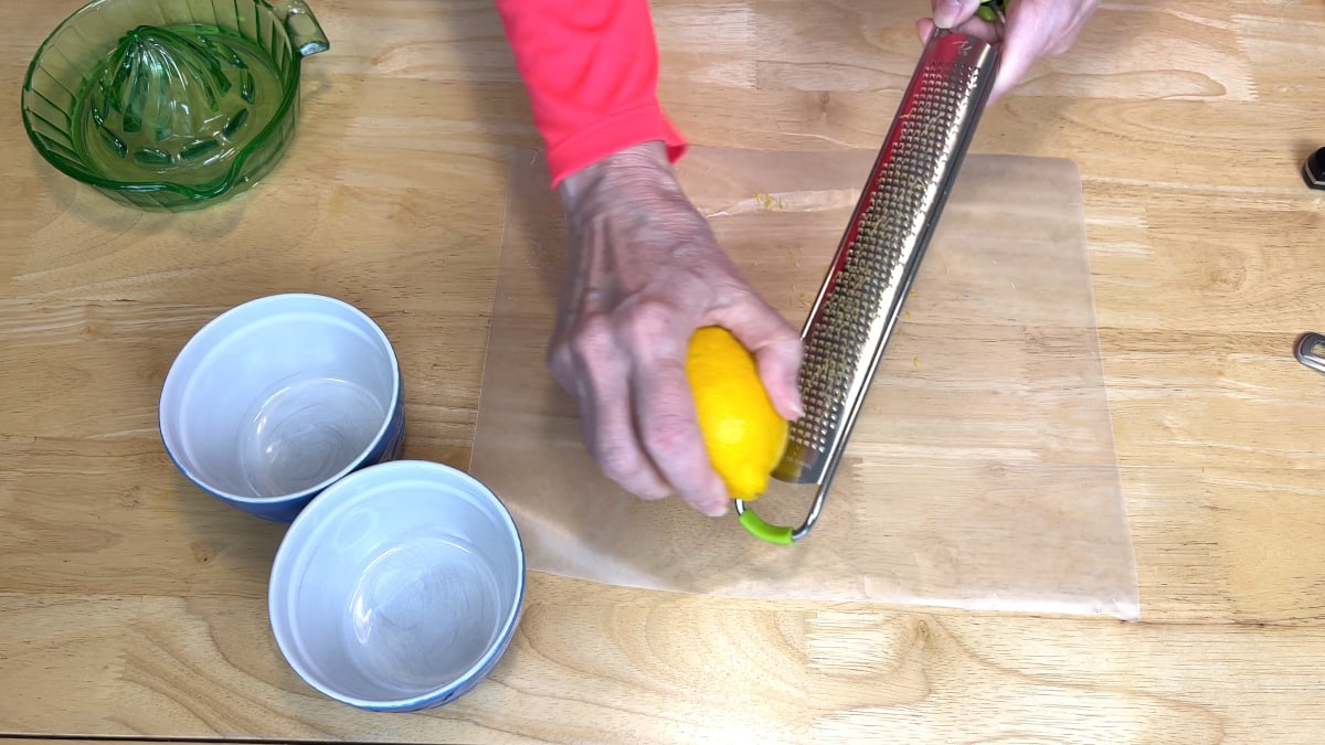 A person zests a lemon with a grater over parchment paper on a wooden table, with two empty bowls and a green citrus juicer nearby.