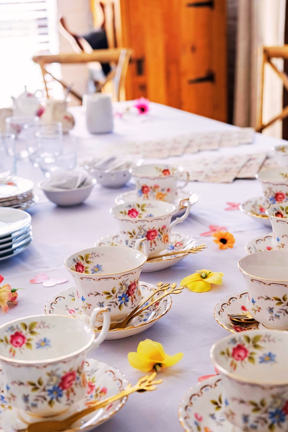 A table set with floral teacups, saucers, and gold utensils on a white tablecloth, decorated with scattered artificial flowers and elegant bridal shower desserts.