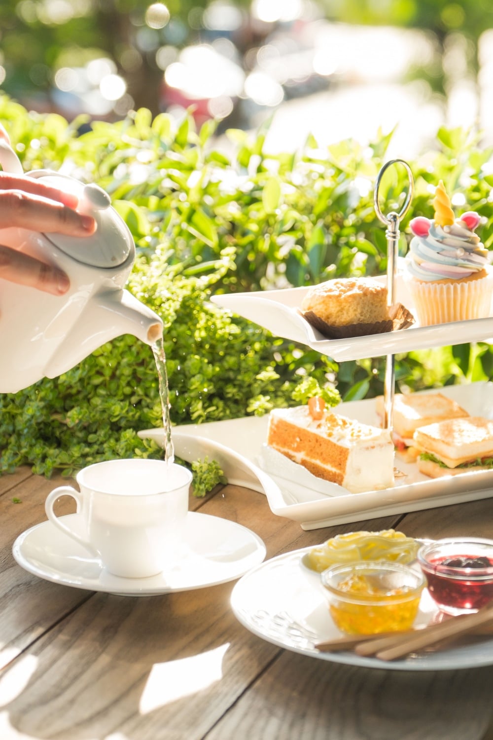 A hand pours tea from a teapot into a cup beside a two-tier tray filled with bridal shower desserts, including pastries, sandwiches, a cupcake, and small dishes of jam on a wooden table outdoors.