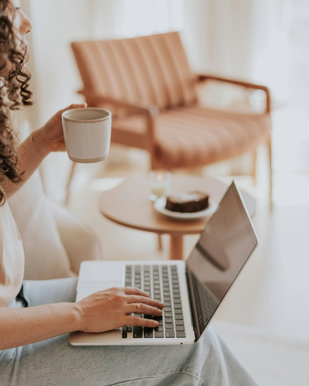 A person sits on a couch using a laptop, holding a coffee mug, with a small table and a dessert in the background.