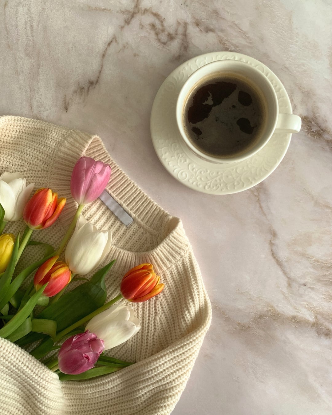 A cup of black coffee sits on a white saucer next to a cream-colored sweater holding a bouquet of tulips on a marble surface.