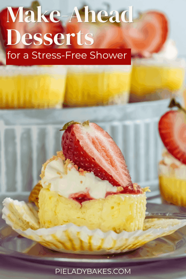A mini cheesecake topped with whipped cream and a fresh strawberry sits on a cupcake liner, with more desserts and strawberries in the background.