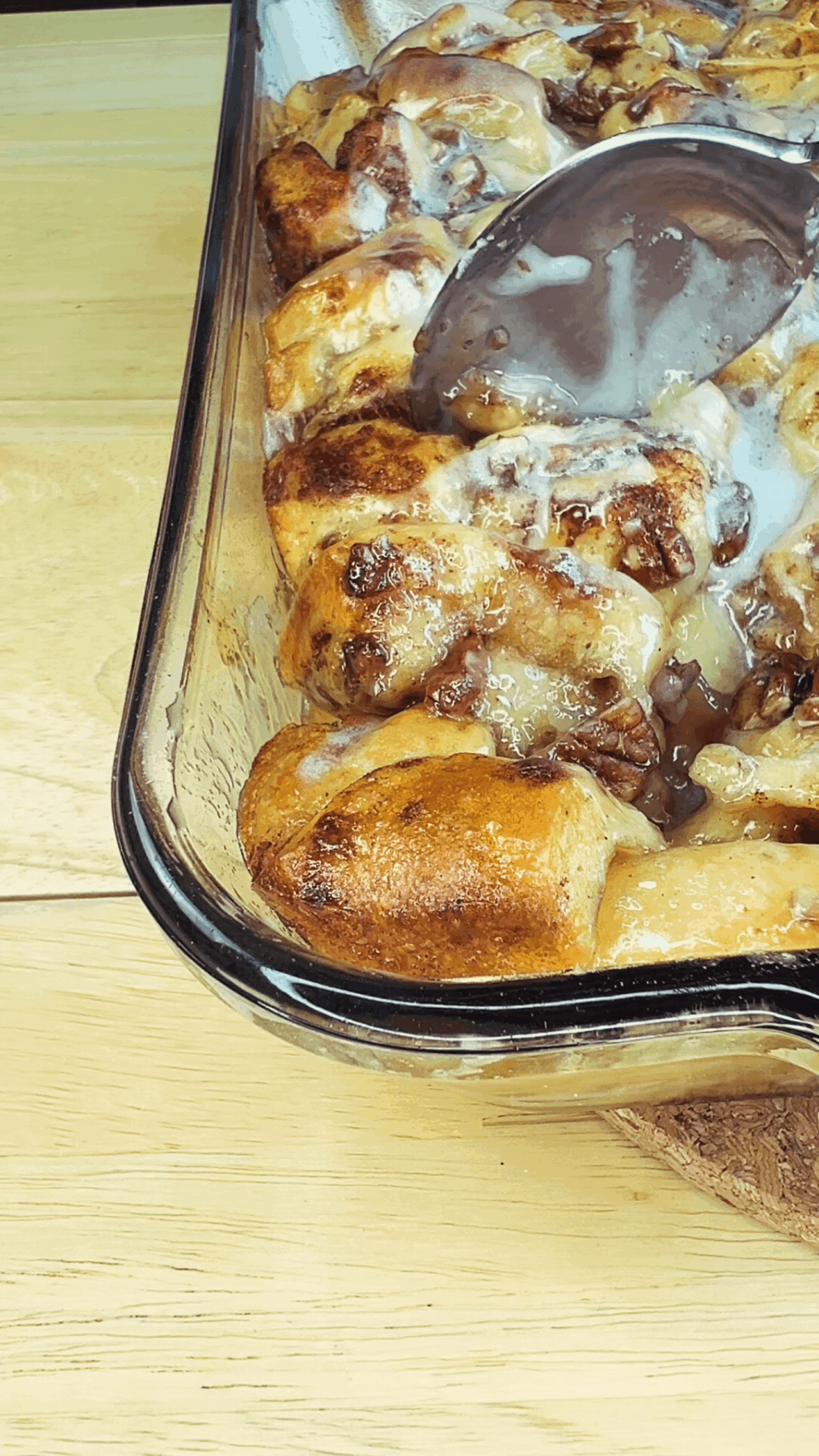 A glass baking dish containing cinnamon rolls topped with icing, with a spatula partially lifting one roll.