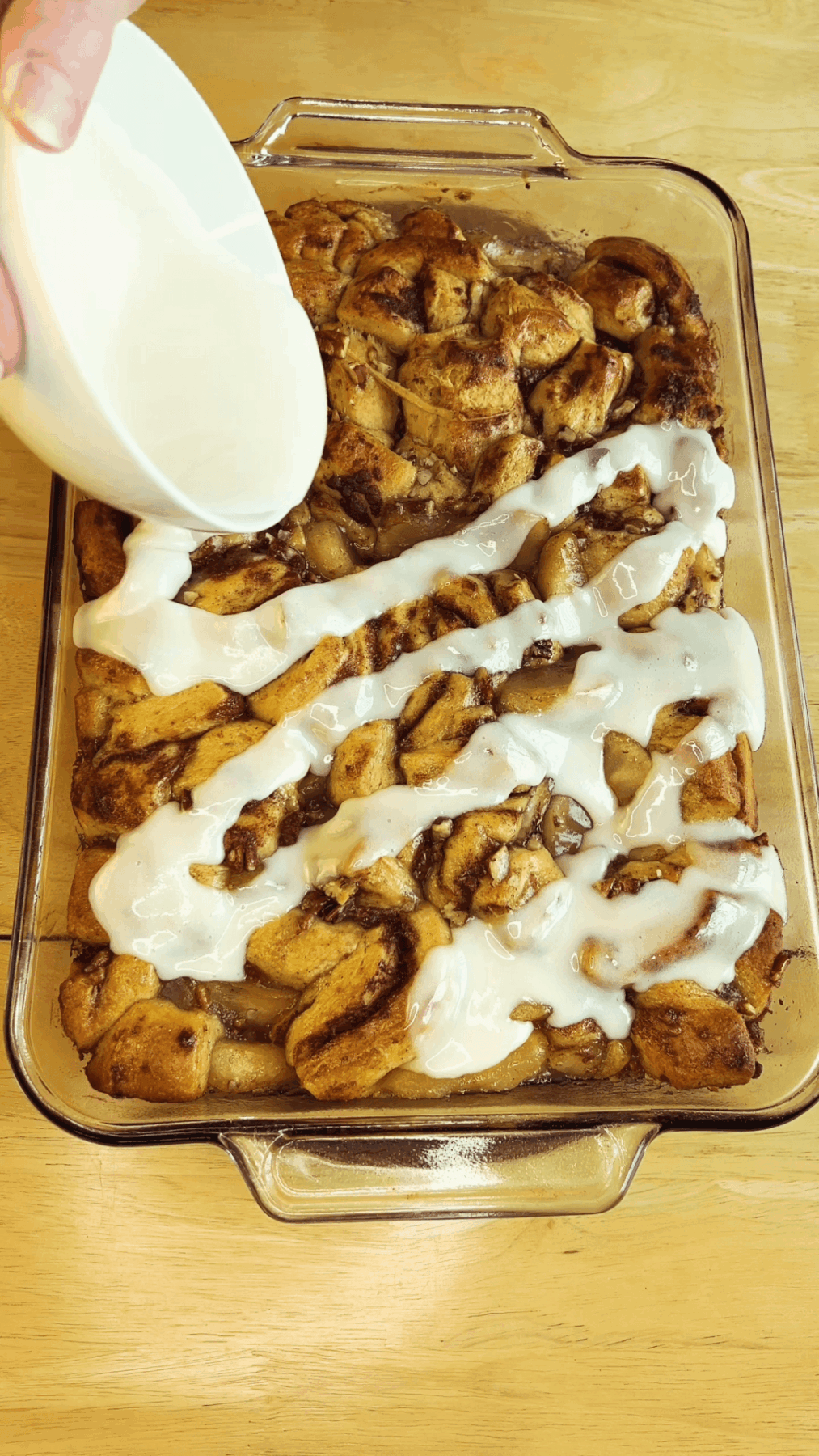 A hand pours white icing over a tray of freshly baked cinnamon roll desserts on a wooden table.