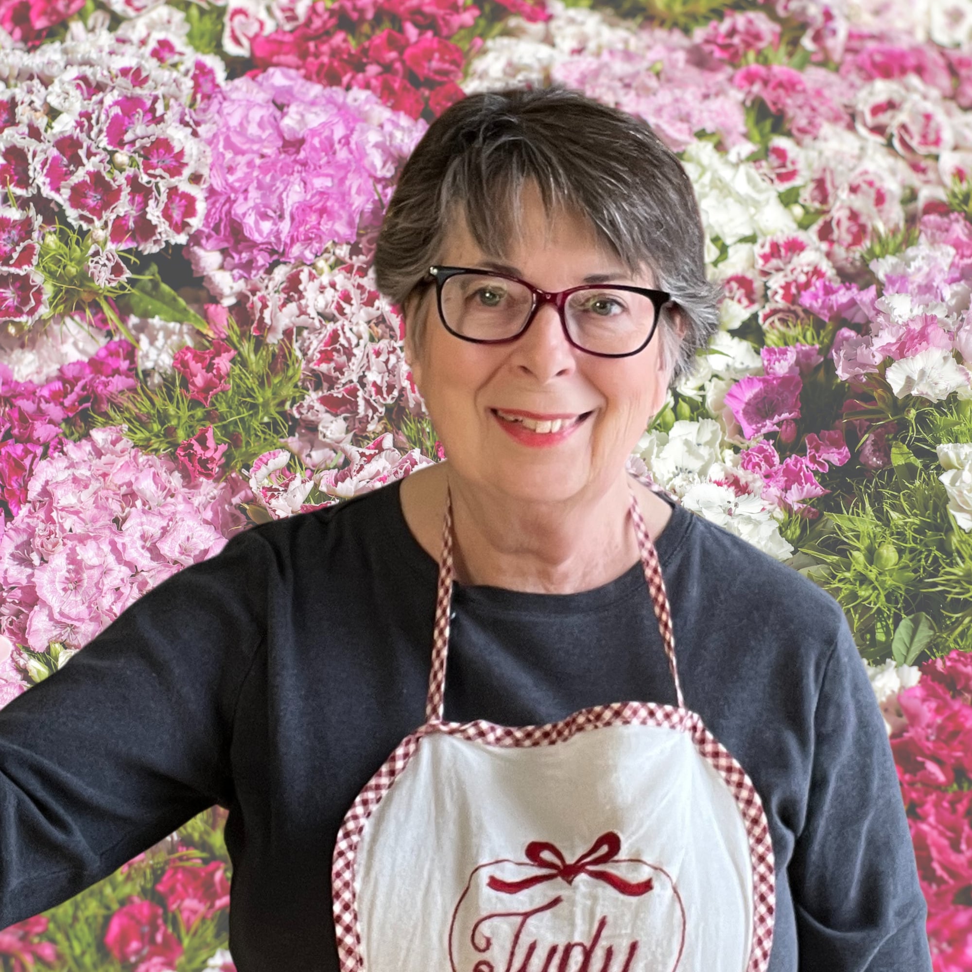 An older woman with short gray hair and glasses smiles, wearing a dark shirt and a checkered apron that says "Judy," standing in front of a background of pink and white flowers.