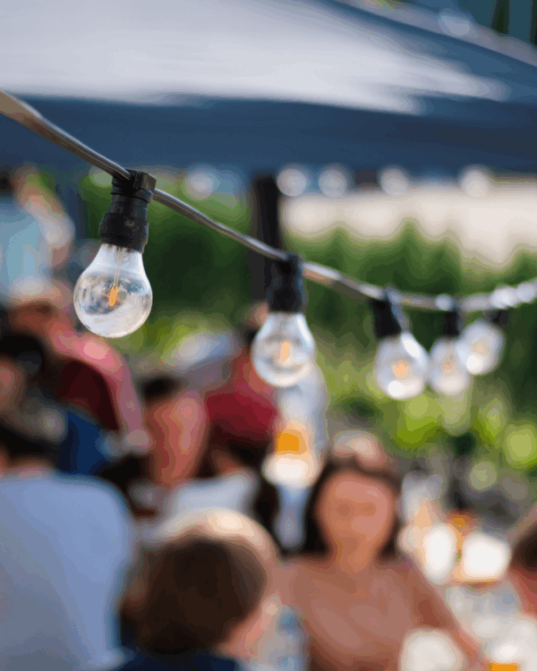 A strand of hanging light bulbs is in focus, with people sitting and talking at blurred outdoor tables in the background.