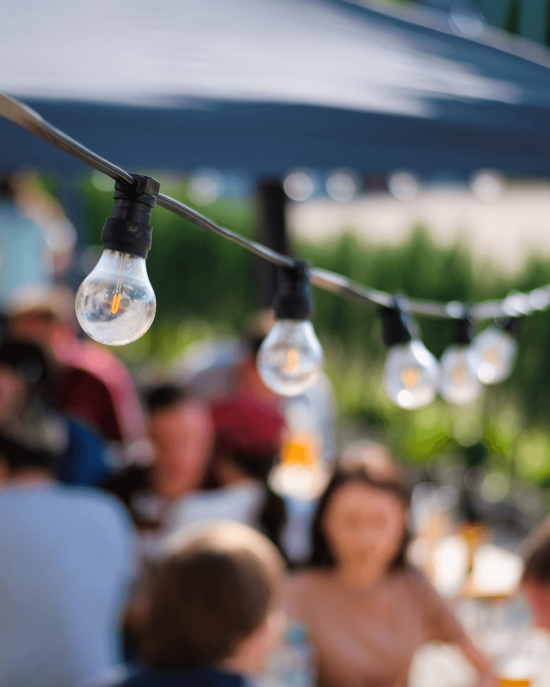 A strand of hanging light bulbs is in focus, with people sitting and talking at blurred outdoor tables in the background.
