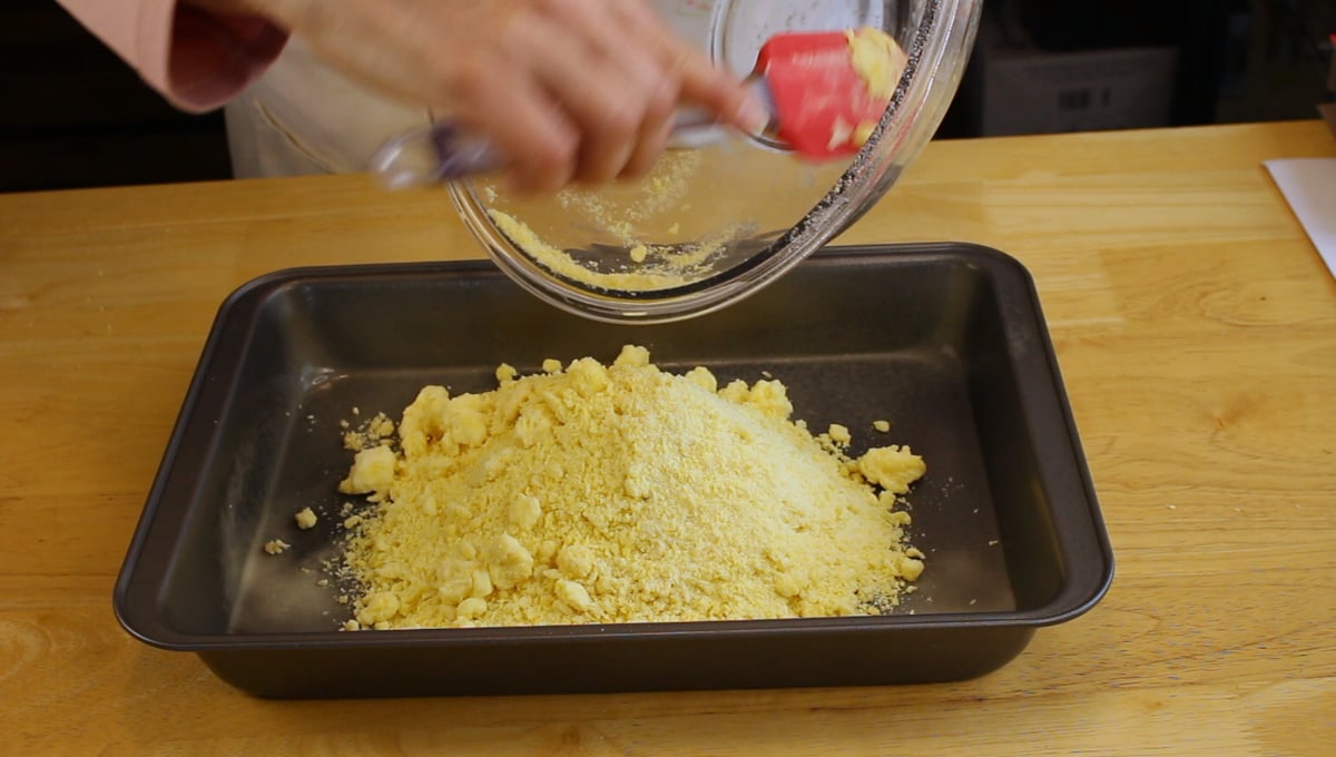 A person pours yellow crumbly dough mixture from a glass bowl into a rectangular baking pan on a wooden surface.