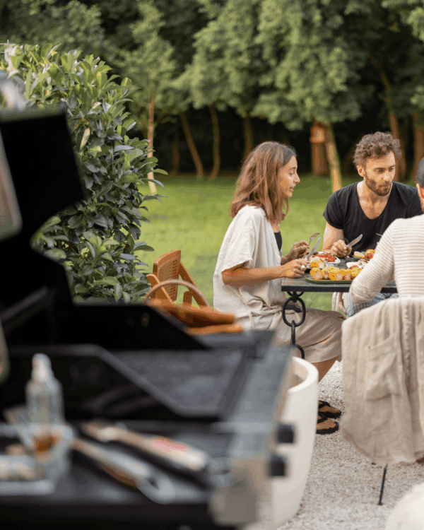 Three people sit at an outdoor table eating a meal together, with a barbecue grill and utensils visible in the foreground. Trees and greenery are in the background.