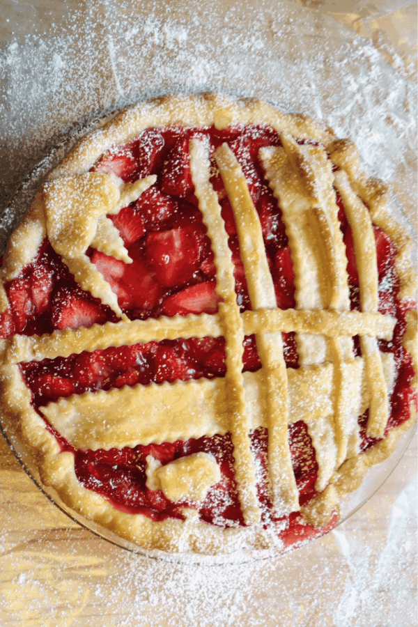 A strawberry pie with a lattice crust and decorative pastry shapes, dusted with powdered sugar, on a lightly floured surface.