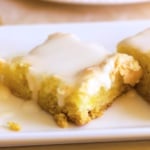 Three rectangular pieces of lemon cake mix cheesecake bars are arranged on a white plate, each topped with a glossy layer of white icing.