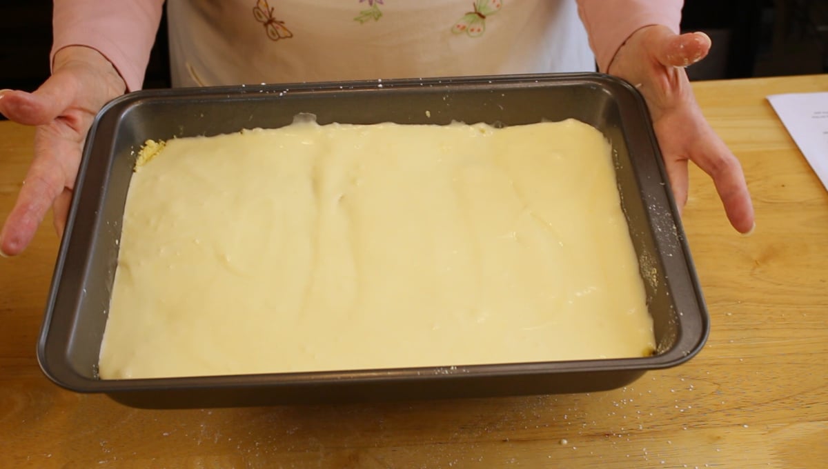 A person holding a baking pan filled with an unbaked layer of batter, placed on a wooden table.
