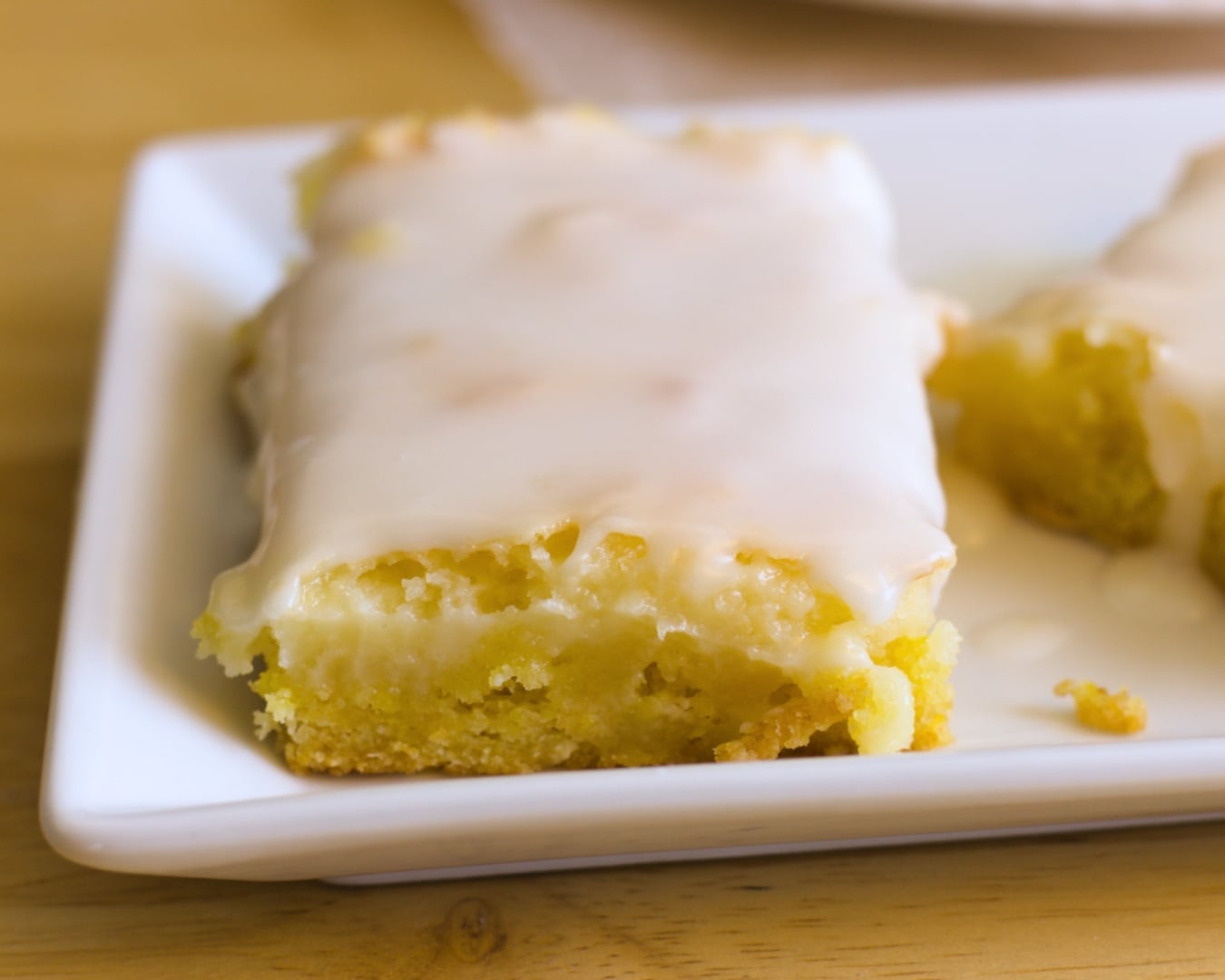 A rectangular slice of yellow sheet cake made with lemon cake mix cheesecake bars, topped with a thick layer of white icing and served on a white plate.