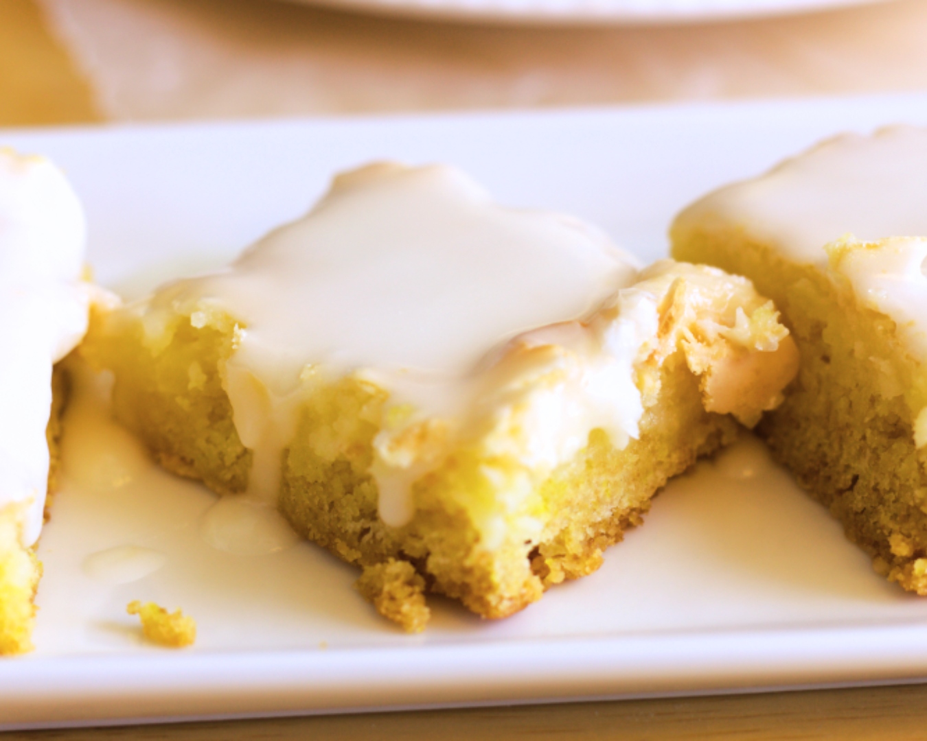 Three rectangular pieces of lemon cake mix cheesecake bars are arranged on a white plate, each topped with a glossy layer of white icing.
