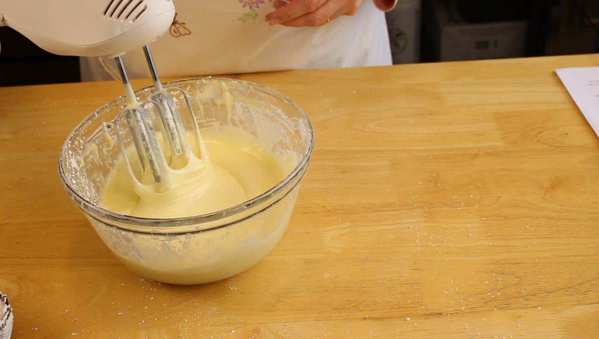 A person uses an electric hand mixer to blend creamy batter in a glass bowl on a wooden table.