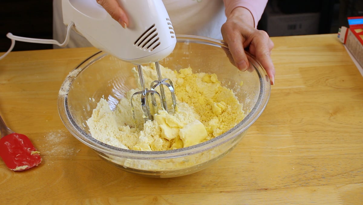 A person uses an electric hand mixer to blend butter and flour in a glass bowl on a wooden table.