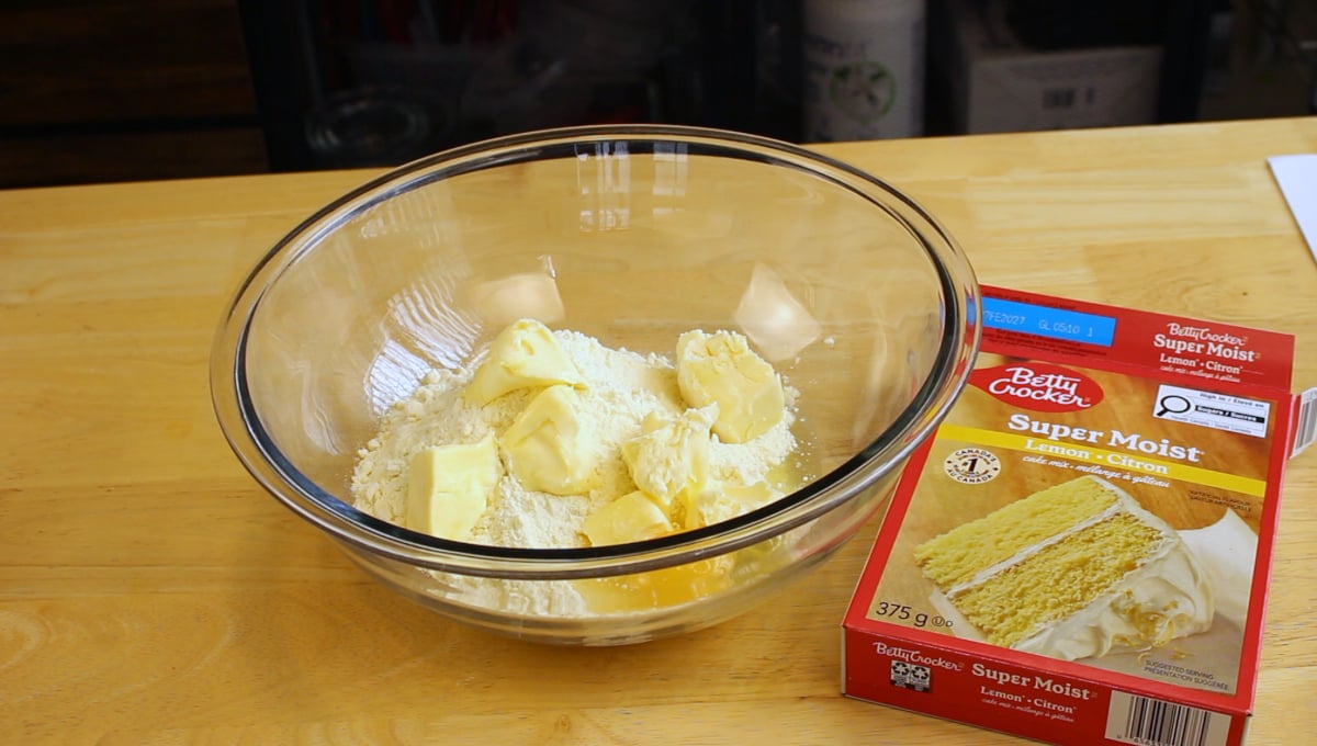 A glass bowl with flour and chunks of butter on a wooden table, next to a box of Betty Crocker Super Moist lemon cake mix.