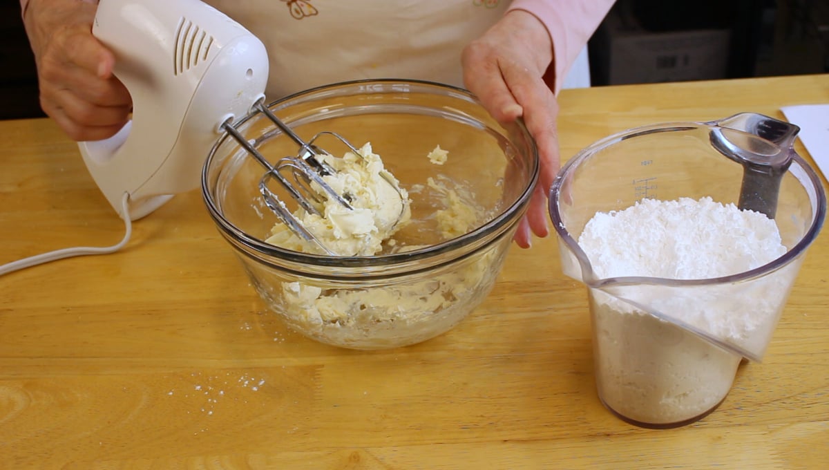 A person uses a hand mixer to blend ingredients in a glass bowl, with a measuring cup of powdered sugar nearby on a wooden table.