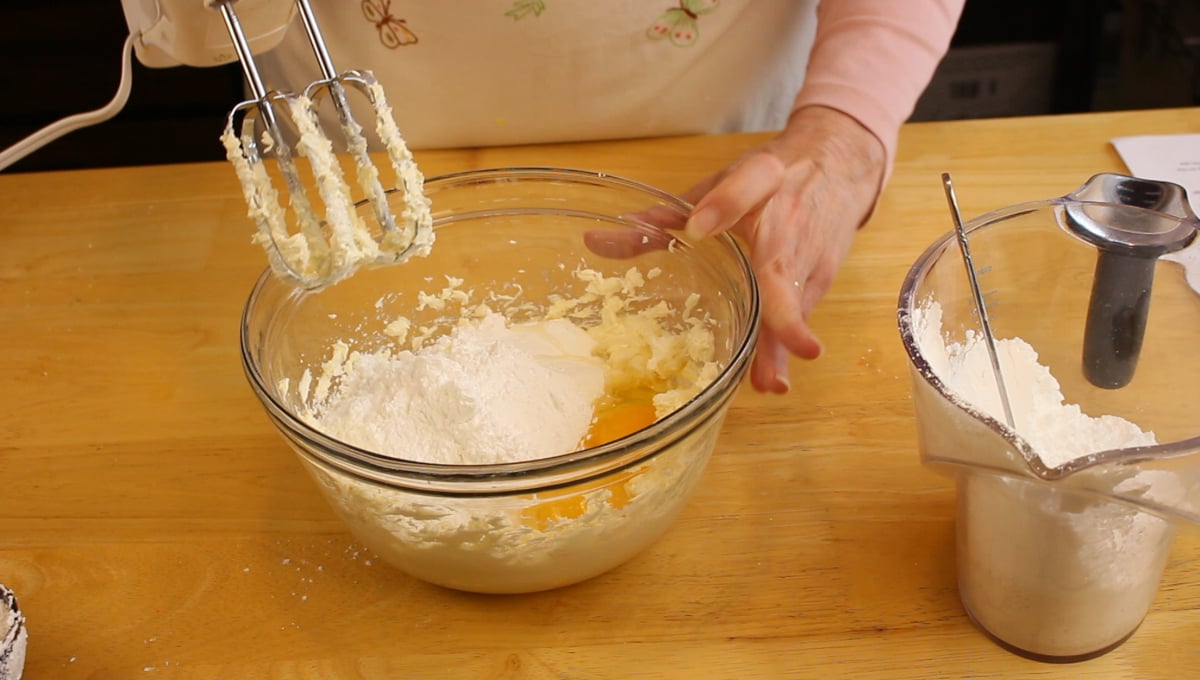 Person using a hand mixer to blend flour, eggs, and other ingredients in a glass bowl on a wooden surface, with extra flour in a measuring jug nearby.