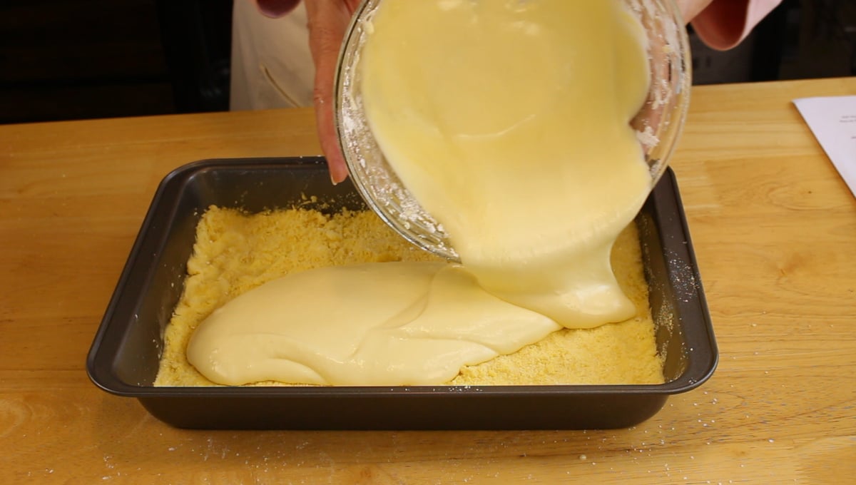 A person pours creamy batter from a glass bowl into a square baking pan with a crumbly crust on a wooden table.