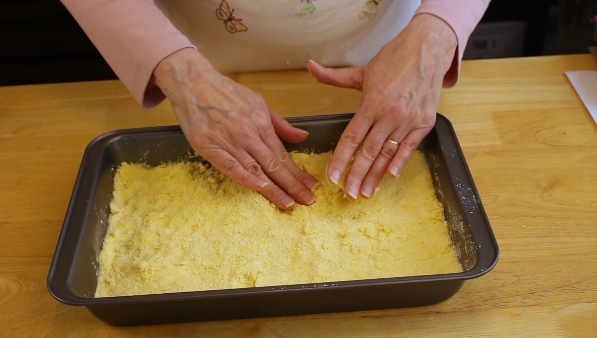 A person presses a crumbly yellow mixture into a rectangular baking pan on a wooden surface.