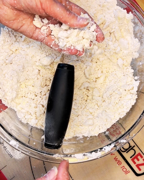 A hand holds a clump of crumbly dough mixture over a bowl with a pastry blender inside, preparing ingredients for baking.