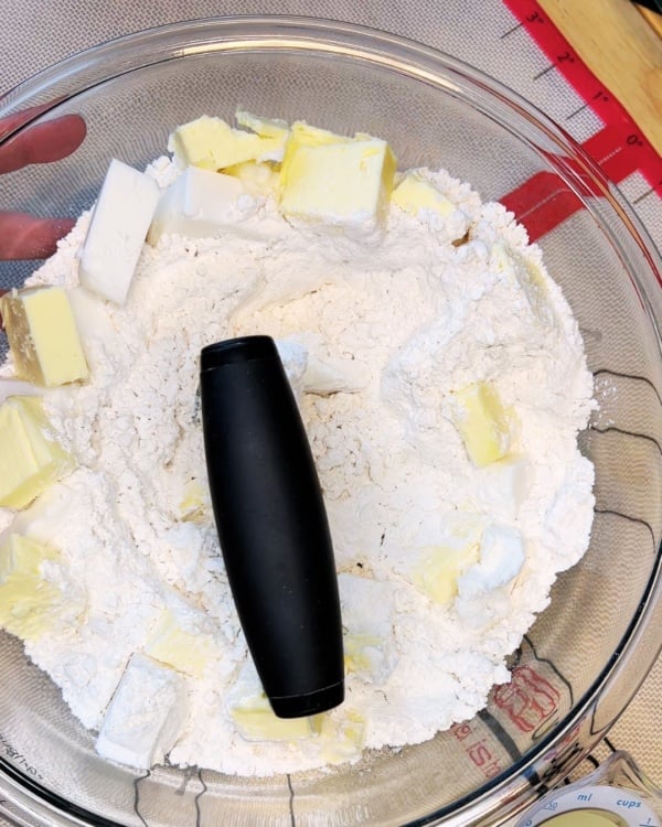 A glass bowl with flour and cubed butter, a pastry blender placed on top, ready for mixing on a countertop with a red ruler nearby.