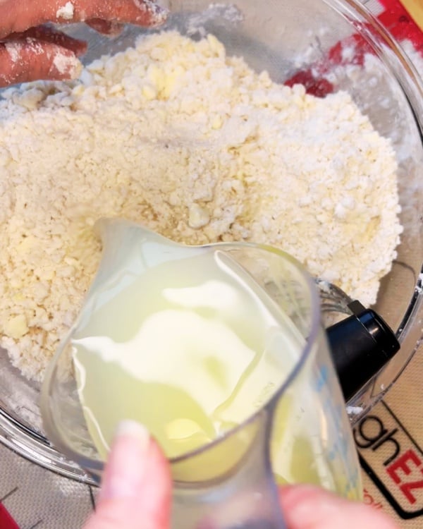 A hand pours liquid from a measuring cup into a mixing bowl filled with flour and butter mixture.