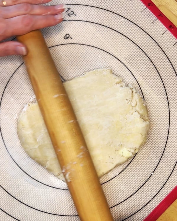 A hand uses a wooden rolling pin to flatten dough on a floured non-stick baking mat with measurement markings.