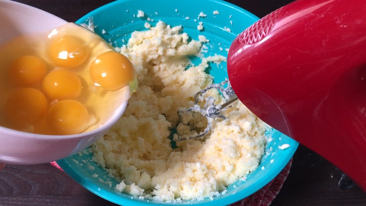 A hand holds a bowl of raw eggs above a blue bowl of creamed butter and sugar, with a red electric mixer nearby.