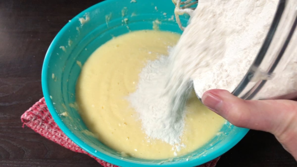 A hand pours flour from a glass bowl into a blue bowl containing yellow batter, with a red cloth underneath the bowl on a dark surface.